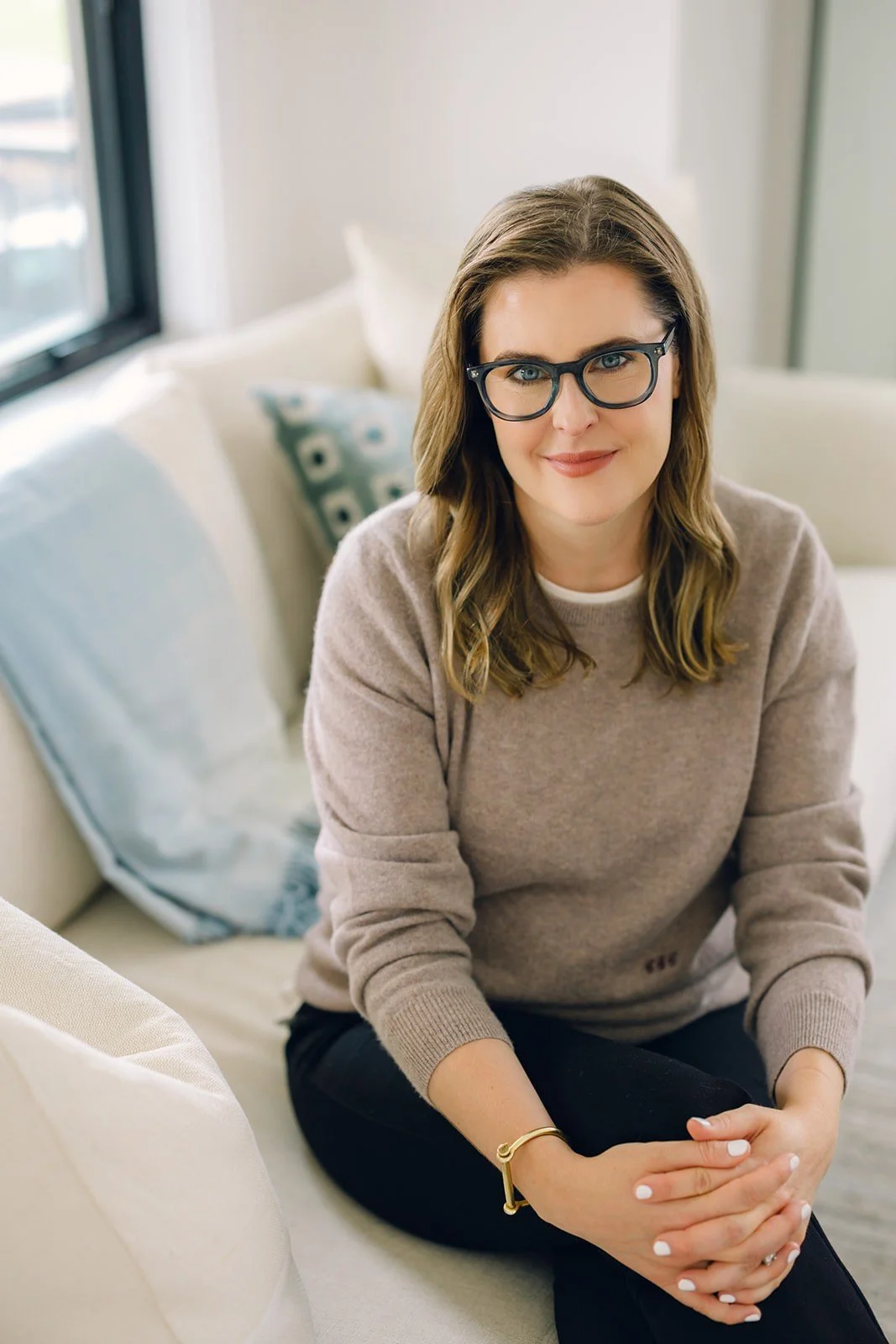 DC-based therapist Claire Connolly wears blue glasses and a beige sweater, sits on a cream couch, smiling empathetically.