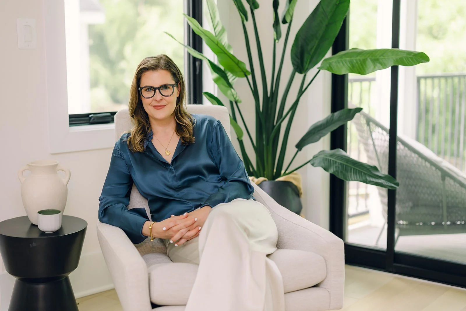 Couples Therapist in Washington DC Claire Connolly is seated in a cream armchair by a window, wearing a blue blouse, with a large green plant in a bright, calming room.