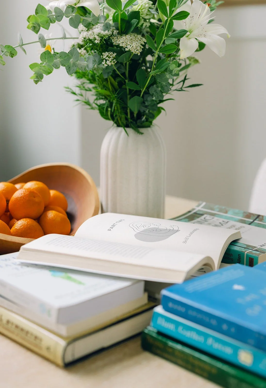 A white vase holding green and white flowers, a bowl of oranges, an open book, and a stack of other books on a table.