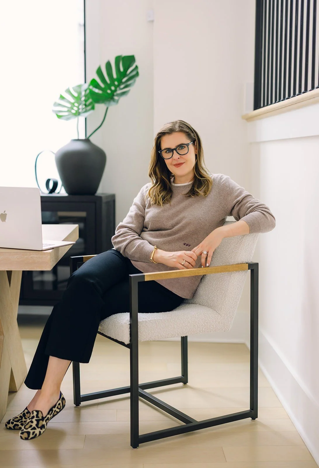 Claire Connolly sits on a modern white chair in a bright room, with a laptop, a table, a large plant, and a black cabinet in the background.