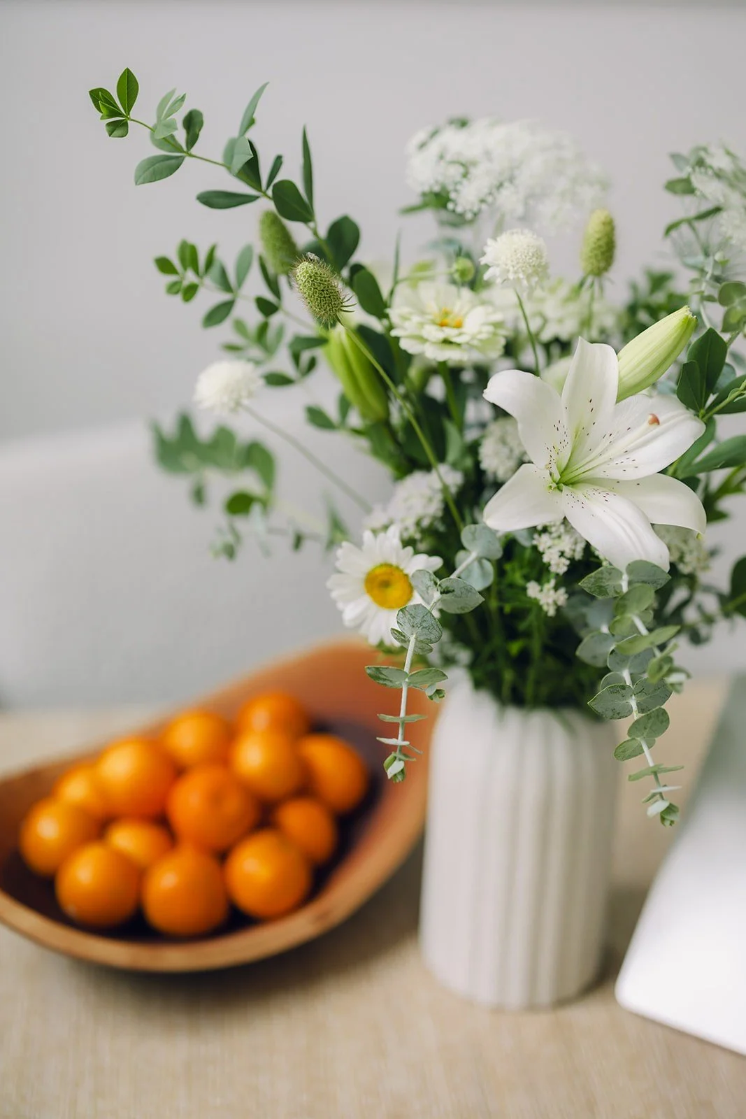 A white vase with a floral arrangement of white lilies, daisies, and greenery, with a bowl of small oranges in the background.