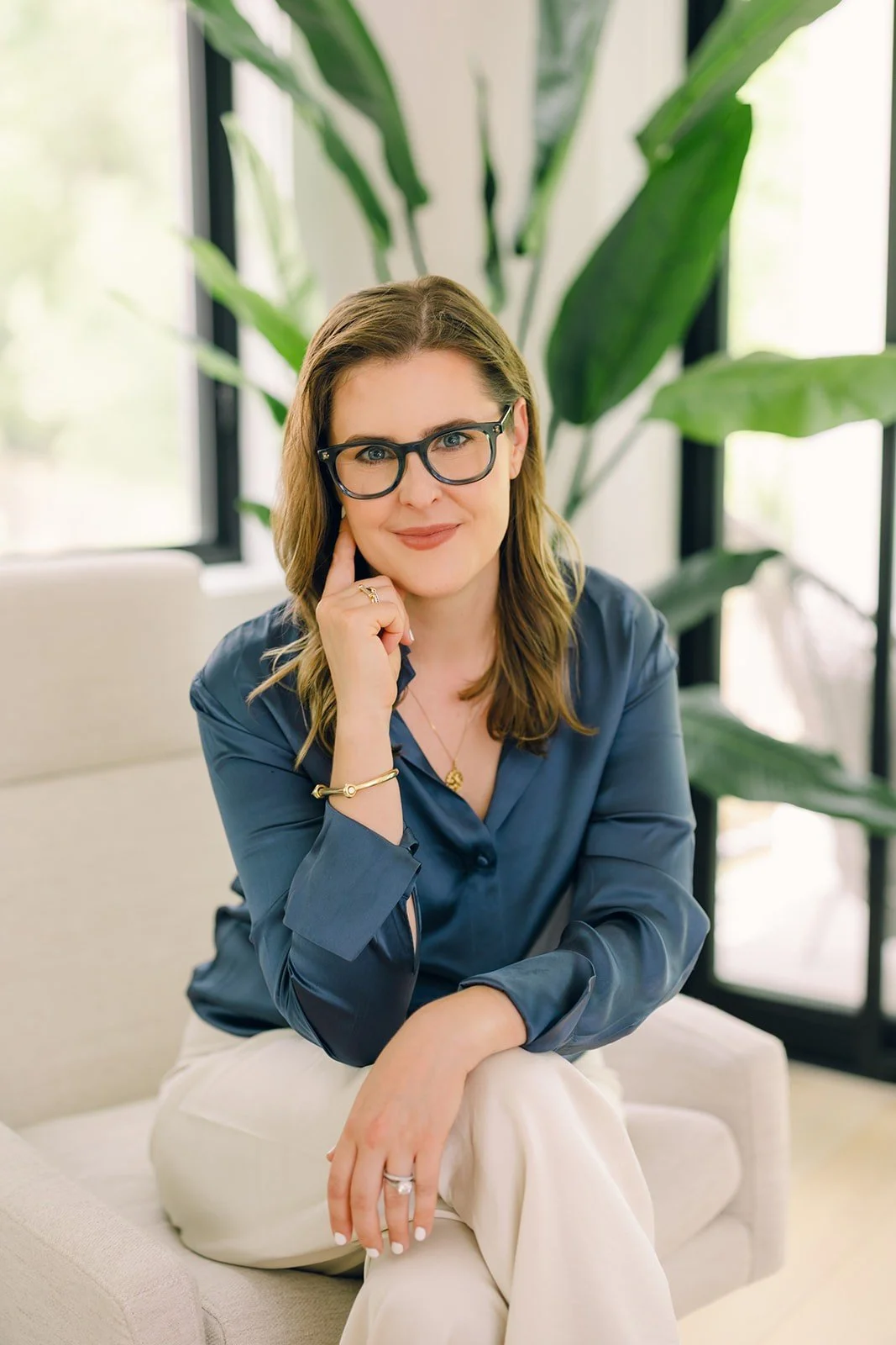 DC-based psychodynamic therapist Claire Connolly rests her chin on her hand, smiling in a blue blouse near large green plants.