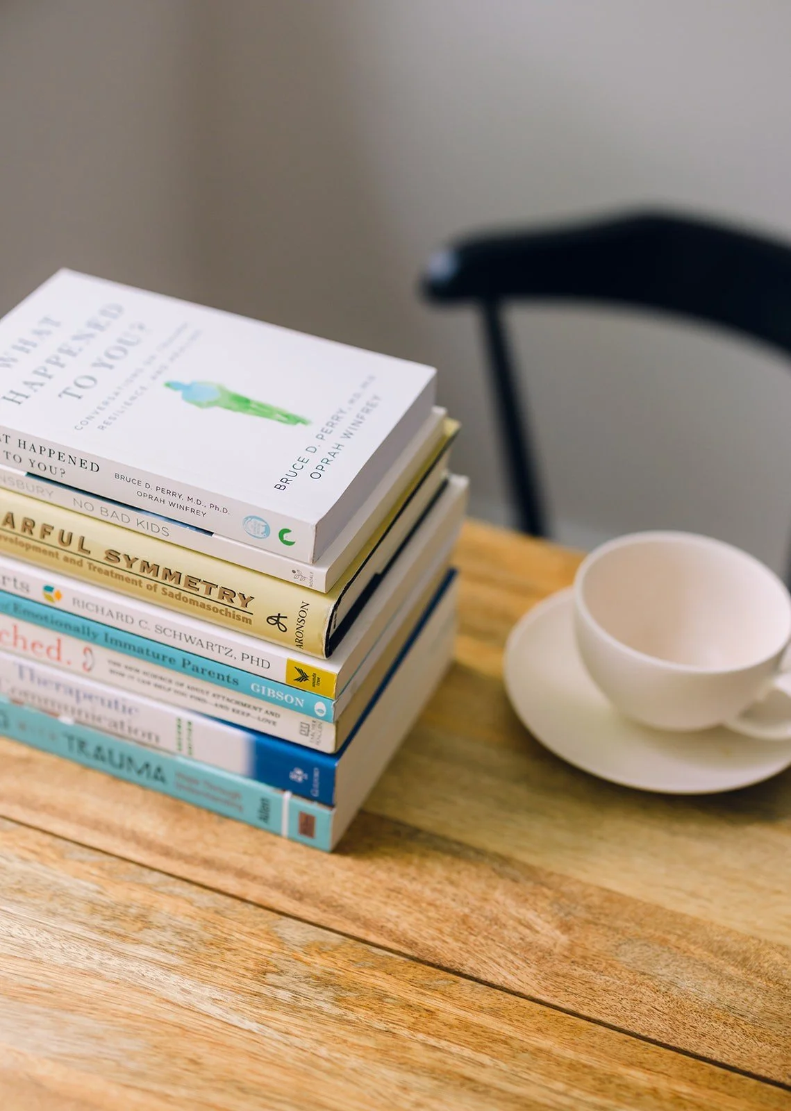 A stack of therapy and psychology books on a wooden table beside a white teacup and saucer, in a calm, warmly lit therapist's office.