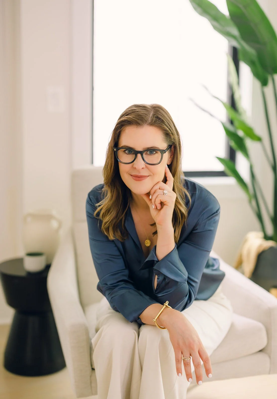 DC therapist Claire Connolly, LICSW, is wearing a blue blouse and glasses, sitting on a cream-colored armchair in a bright room with a large window and green plants, resting her chin on her hand and smiling.