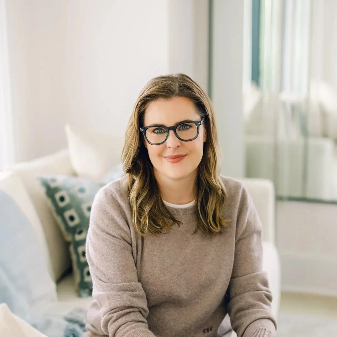 DC therapist Claire Connolly, LICSW is wearing glasses and a beige sweater, and sitting on a white sofa in a bright room with blue decorative pillows and large windows.