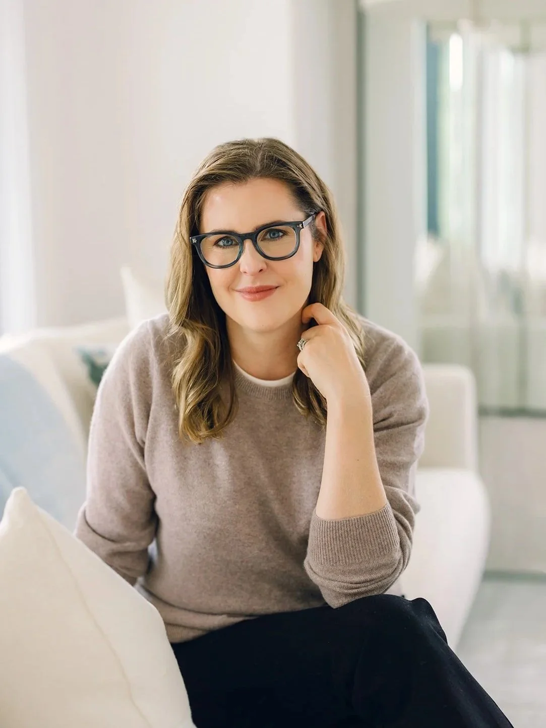 Claire Connolly, LICSW is wearing glasses, a beige sweater, and black pants, sitting on a light-colored sofa in a bright room, smiling at the camera.