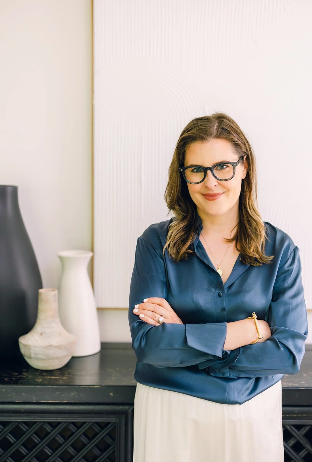 Claire Connolly stands with arms crossed, smiling in a blue blouse, near ceramic vases and framed art in a modern room.