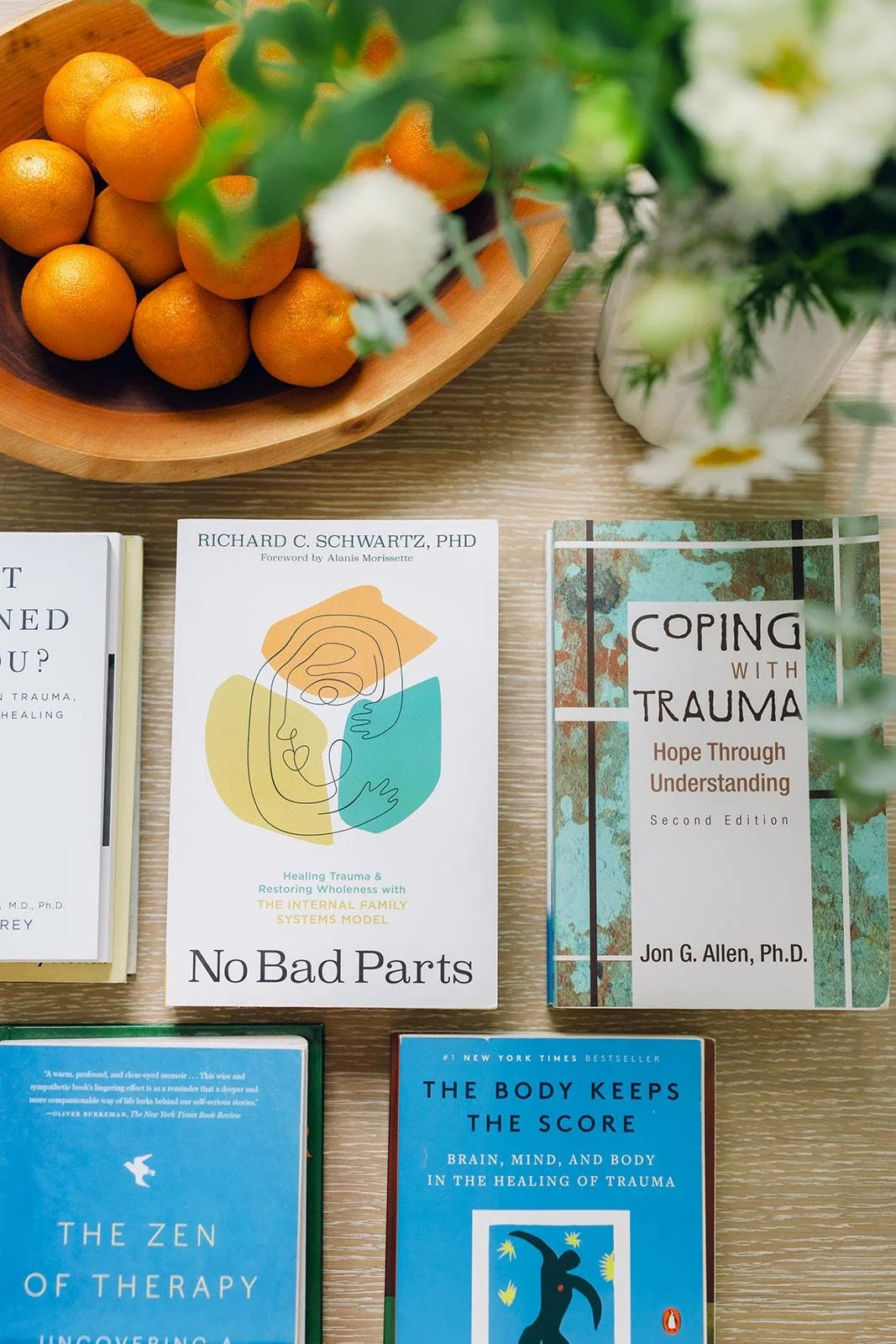 Top of a table with a wooden bowl of oranges, a plant with white flowers, and books related to trauma, therapy, and healing.