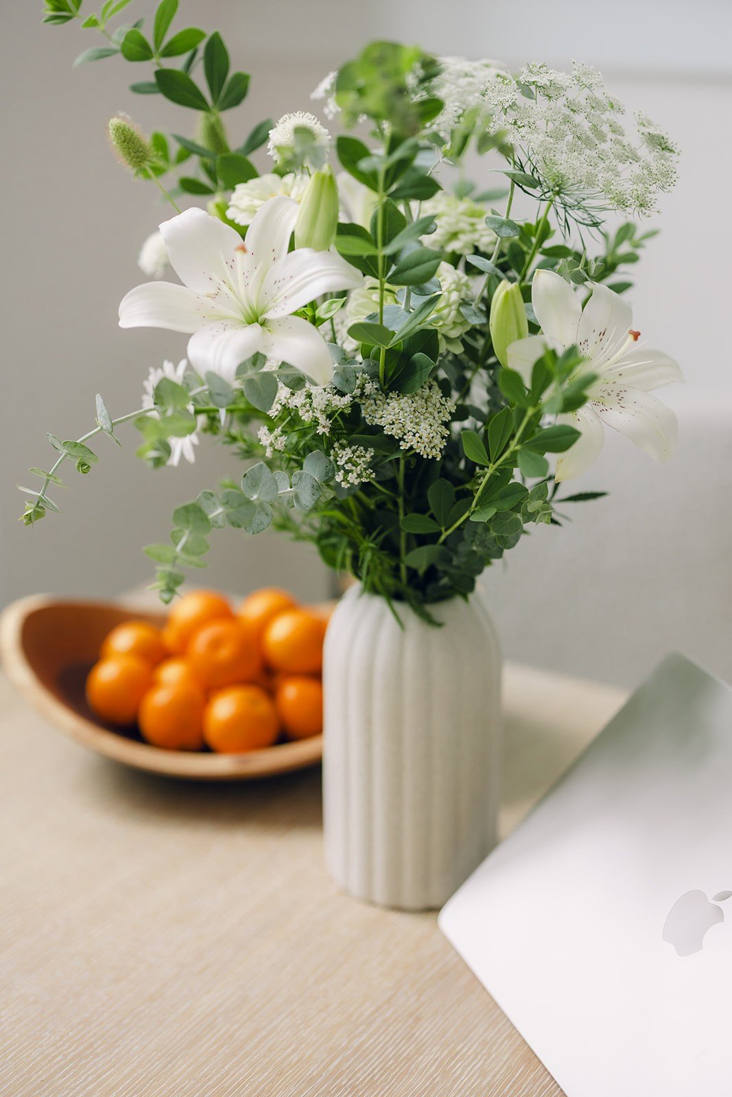A white vase with a bouquet of white lilies, green leaves, and other white flowers on a beige table, with a bowl of oranges and a silver laptop nearby.