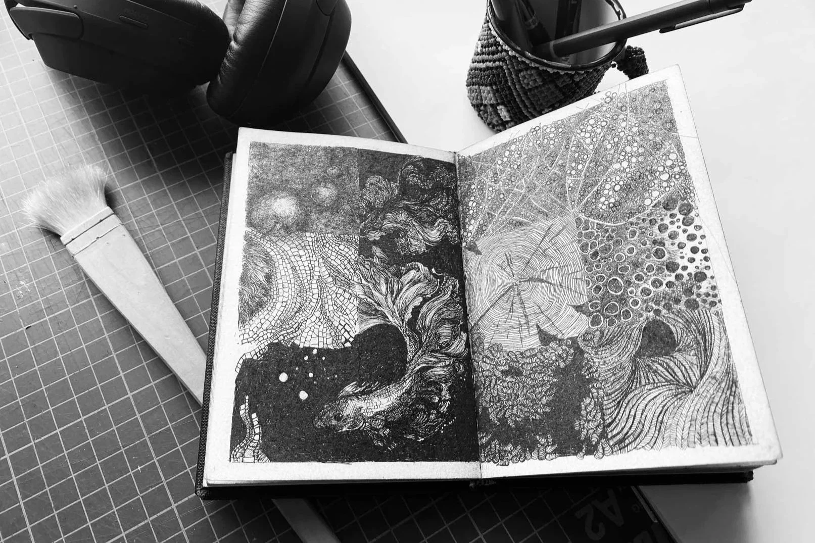 Open sketchbook with intricate black and white line drawings of abstract patterns, placed on a desk with a paintbrush, headphones, and a pencil holder nearby.