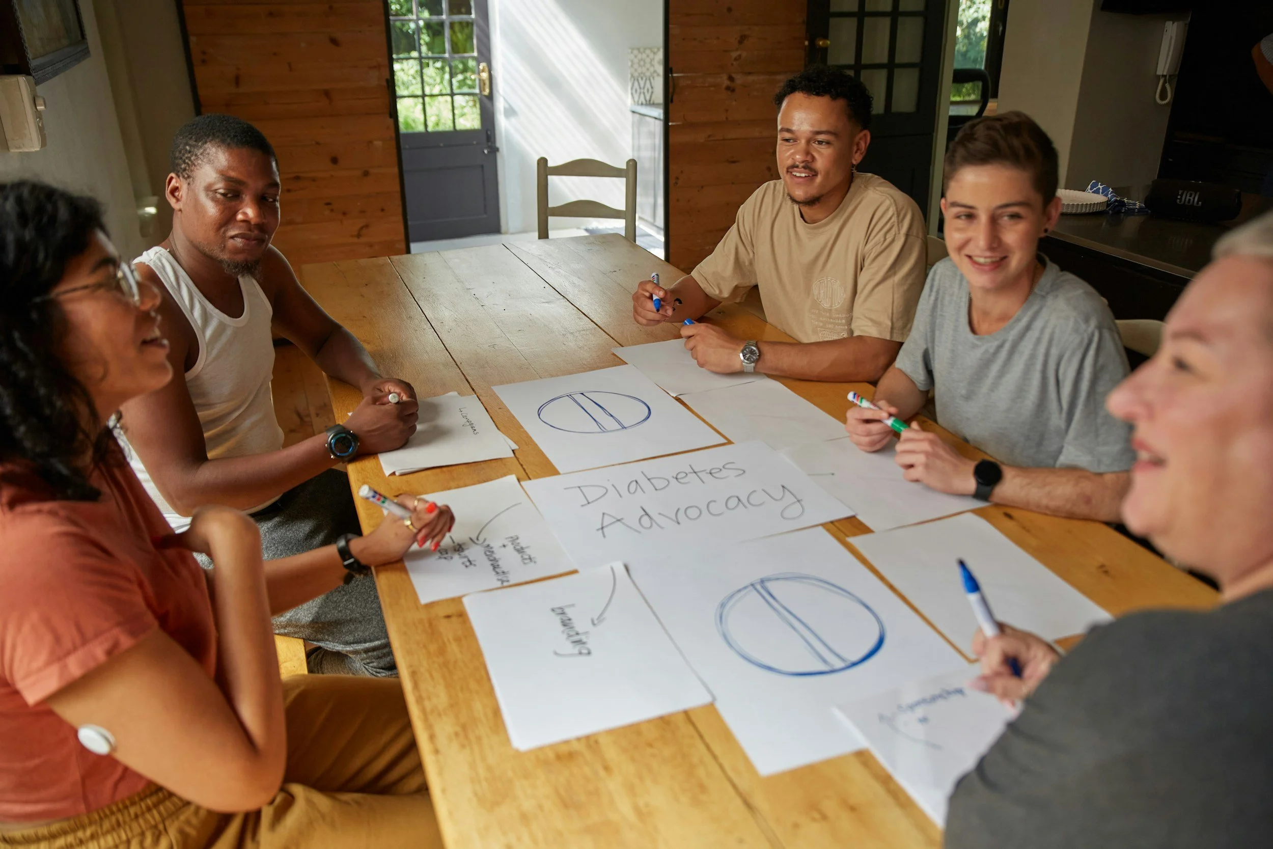 Group of five young adults sitting around a wooden table, engaged in a discussion about diabetes advocacy, with printed charts and notes on the table.