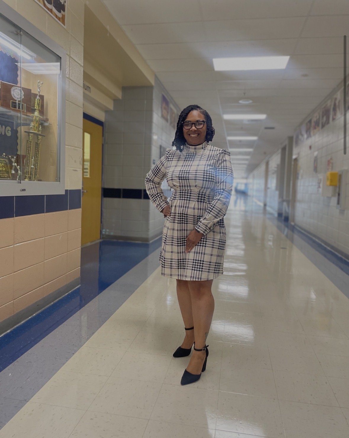 A woman wearing glasses and a plaid dress standing in a school hallway with trophies on display.