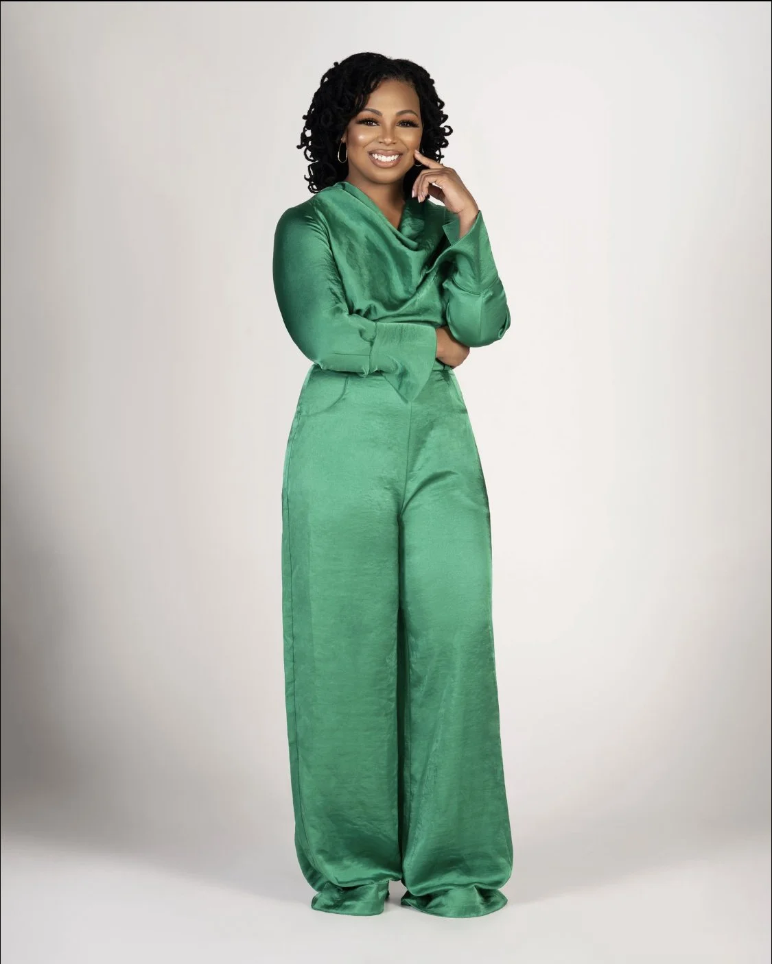 A woman with curly hair wearing a shiny green satin jumpsuit, standing against a white background, smiling and posing with her hand resting on her face.
