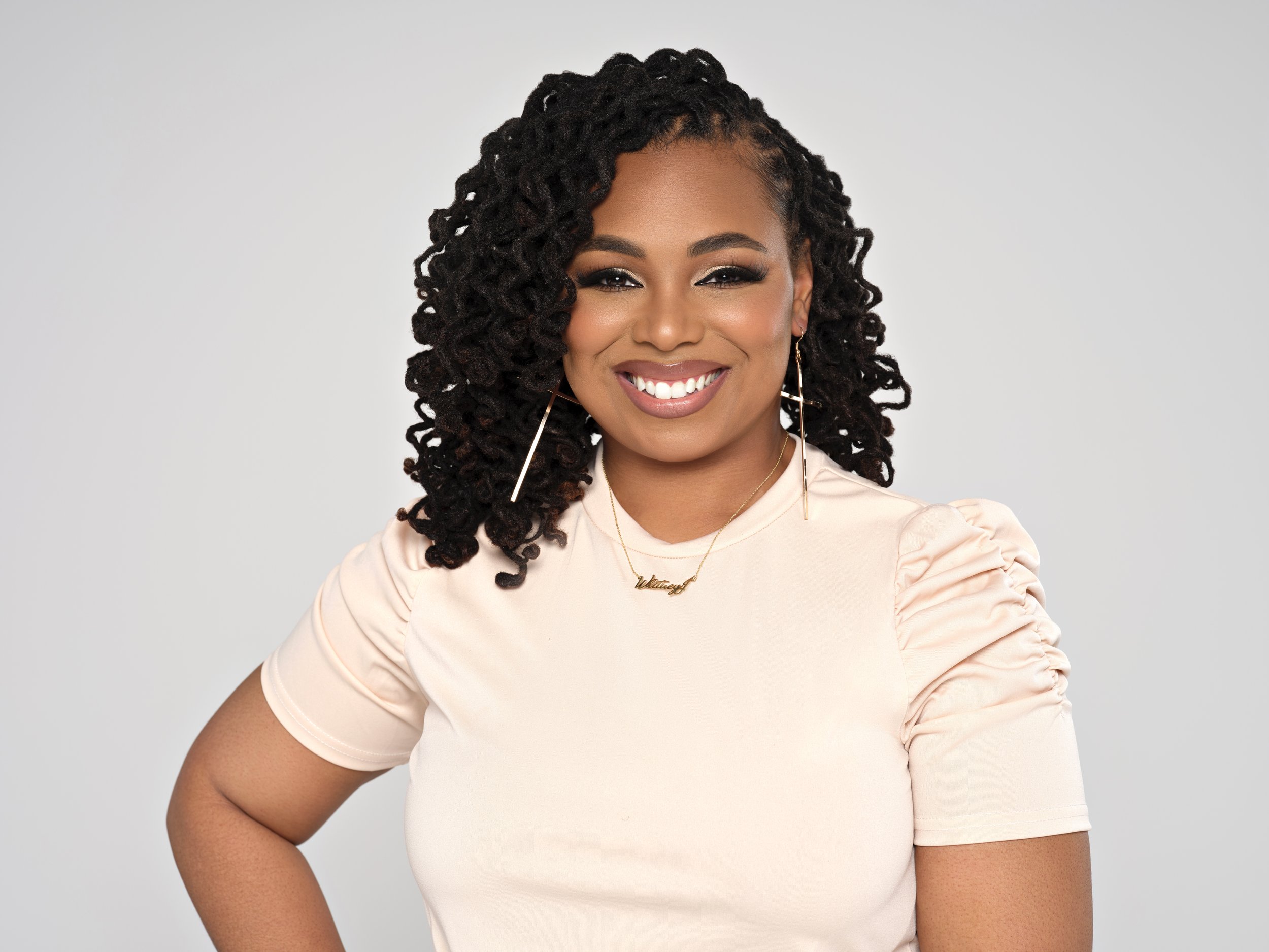 A woman with dark, curly hair styled in twists, smiling, wearing a light-colored top with puffed sleeves, gold earrings, and layered necklaces, standing against a plain light gray background.