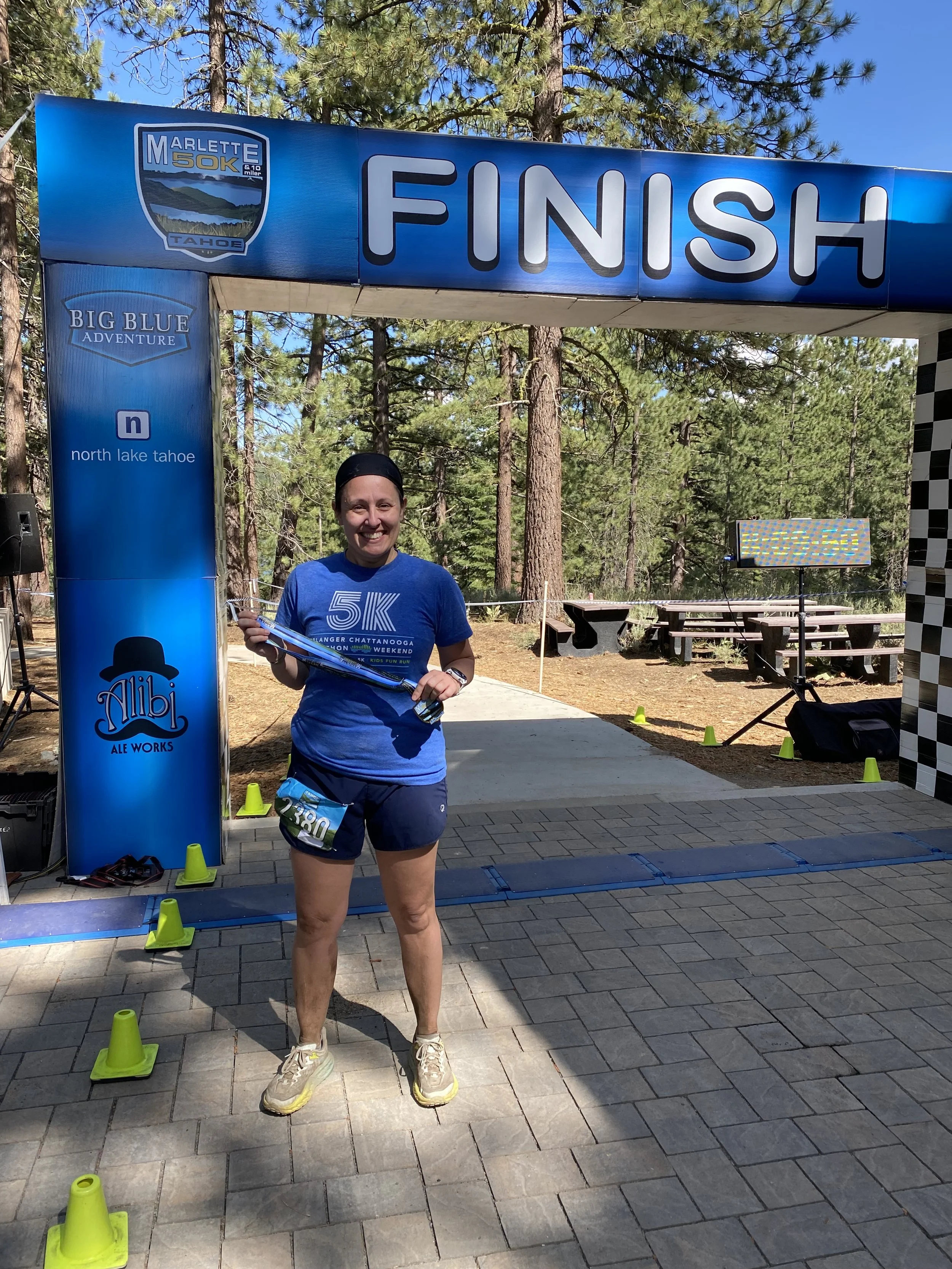 Emily smiling and holding a medal at the finish line of a race event in a wooded outdoor area. She wears a blue race T-shirt, shorts, and running shoes, standing in front of a finish banner with trees in the background.