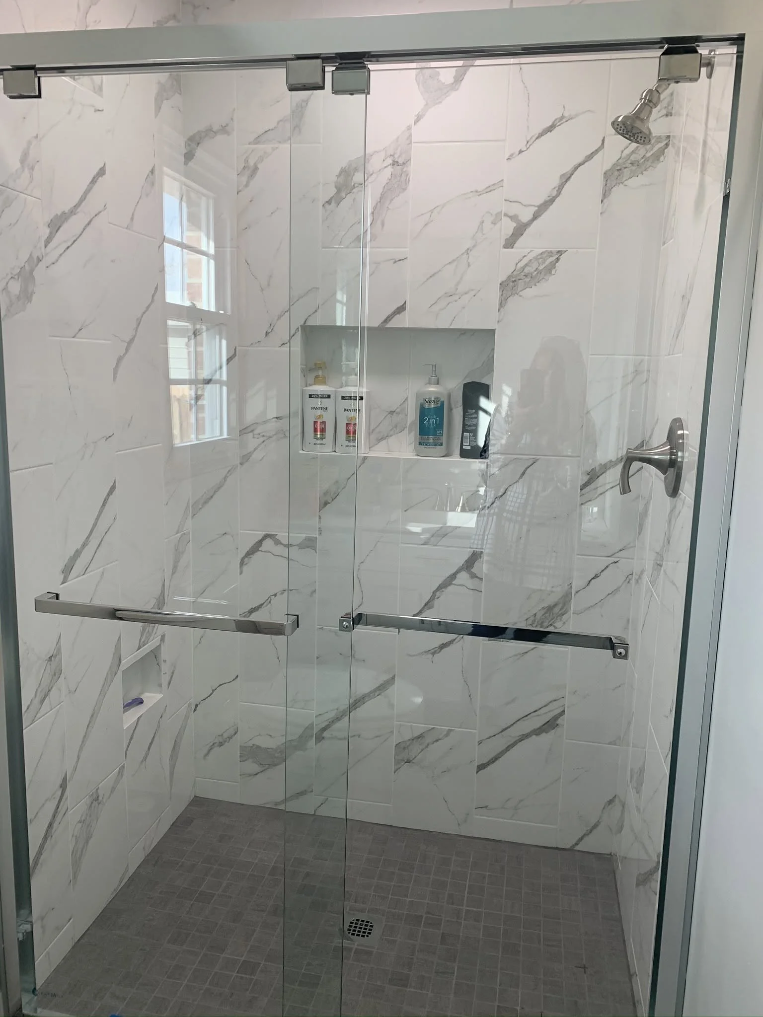 A shower with glass doors, marble walls, and a tiled floor. Inside the shower, there are bottles of shampoo and body wash on a built-in shelf, a showerhead, and a handle for controlling water flow.