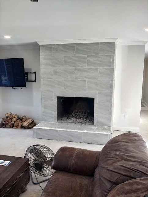 Living room with a gray tiled fireplace, a television mounted on the wall, a portable fan on the floor, a pile of firewood next to the fireplace, and a dark brown leather sofa.
