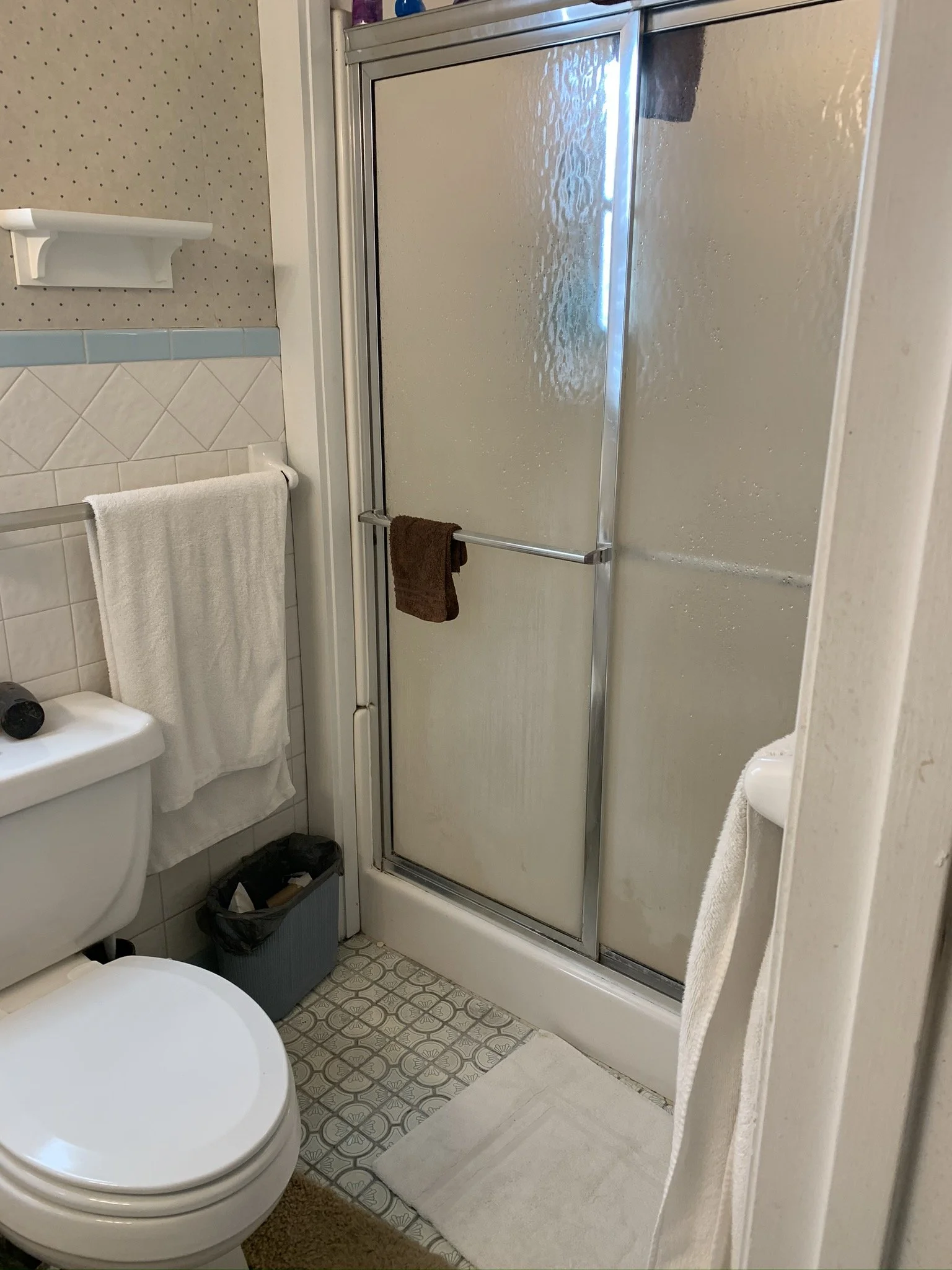 A small bathroom with a toilet, a shower with a frosted glass door, and a white towel hanging on a towel bar. The floor has a patterned tile design.