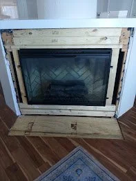 Fireplace installation in progress with a wooden frame surrounding the fireplace opening and a cardboard piece on the floor in front of it.