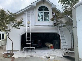 Two workers installing or repairing a garage door on a white house, with ladders on either side and a car inside the garage.