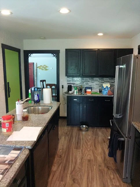 A kitchen with dark cabinets, a stainless steel refrigerator, a granite island counter, and a sliding glass door leading outside. The counter has various cleaning supplies, paper towels, and utensils.
