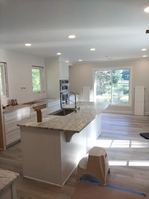 Modern kitchen with white cabinets, marble countertops, stainless steel sink, built-in oven, large window, and sliding glass door leading outside.