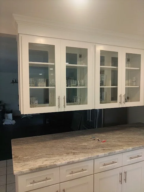 White kitchen upper cabinet with glass doors and visible glassware, white lower cabinets with drawers, and a beige granite countertop.