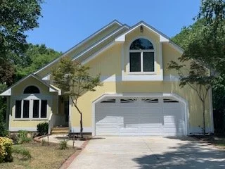 A yellow house with a white garage door, large front window, and an arched window above the garage. There are trees and a driveway in front.