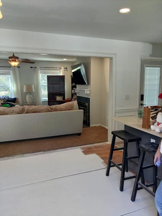 Living room with beige sofa, window, ceiling fan, TV on wall above fireplace, bookshelf, lamp, doorway, and kitchen counter with bar stools.