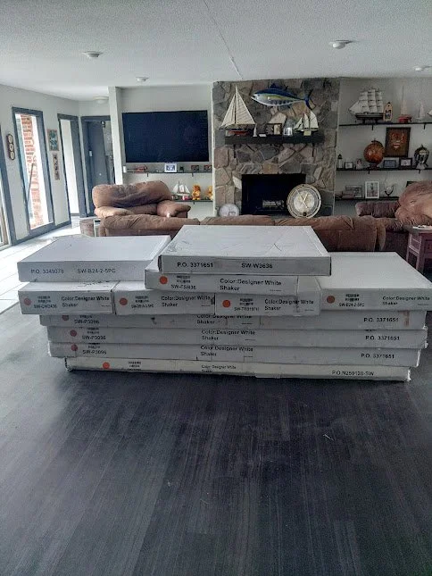 Several boxes of white paint stacked in a living room with a stone fireplace, brown sofas, and a large flat-screen TV on the wall.