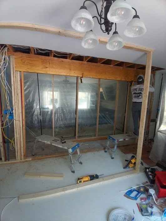 Inside a home under construction, showing a wooden frame wall, work ladders, tools on the floor, and wiring on the left side. A person in a black t-shirt and gray pants stands near the wall.