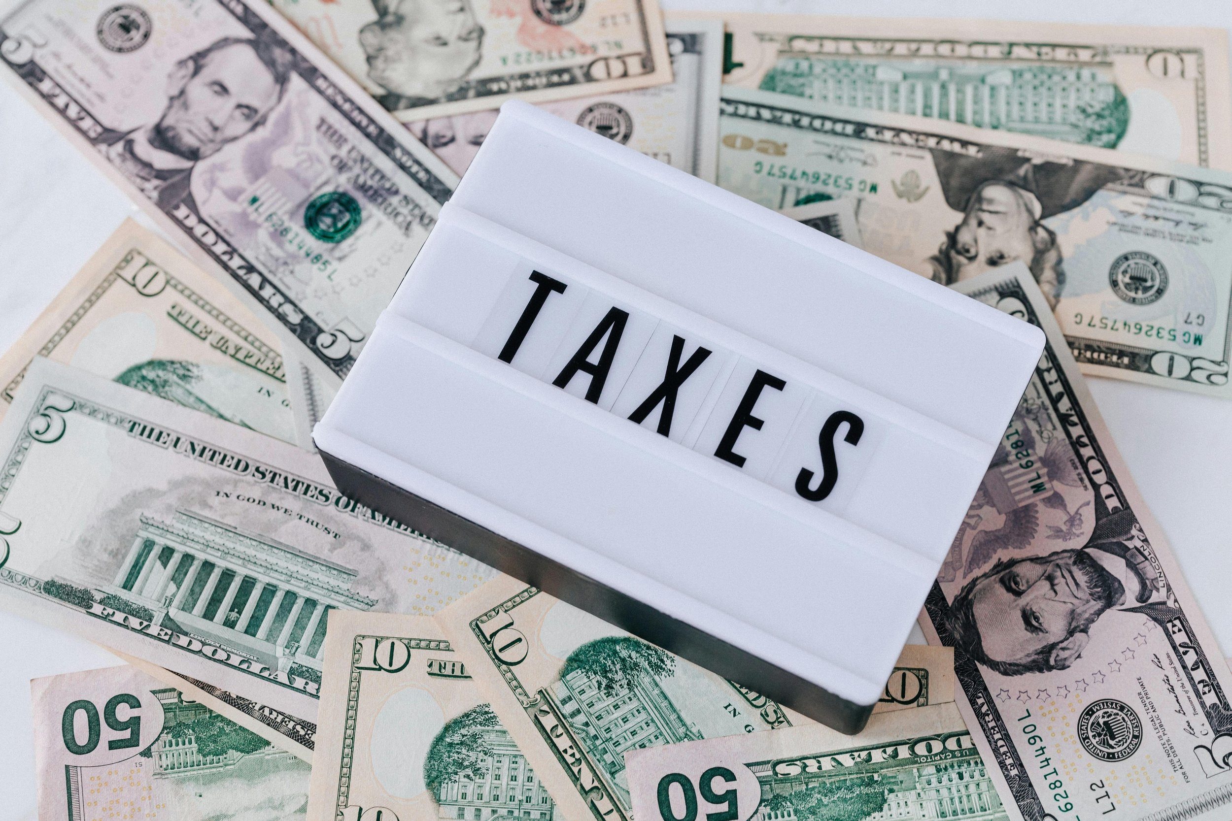 A collection of U.S. fifty-dollar bills and ten-dollar bills scattered around, with a white sign displaying the word 'TAXES' in black letters.