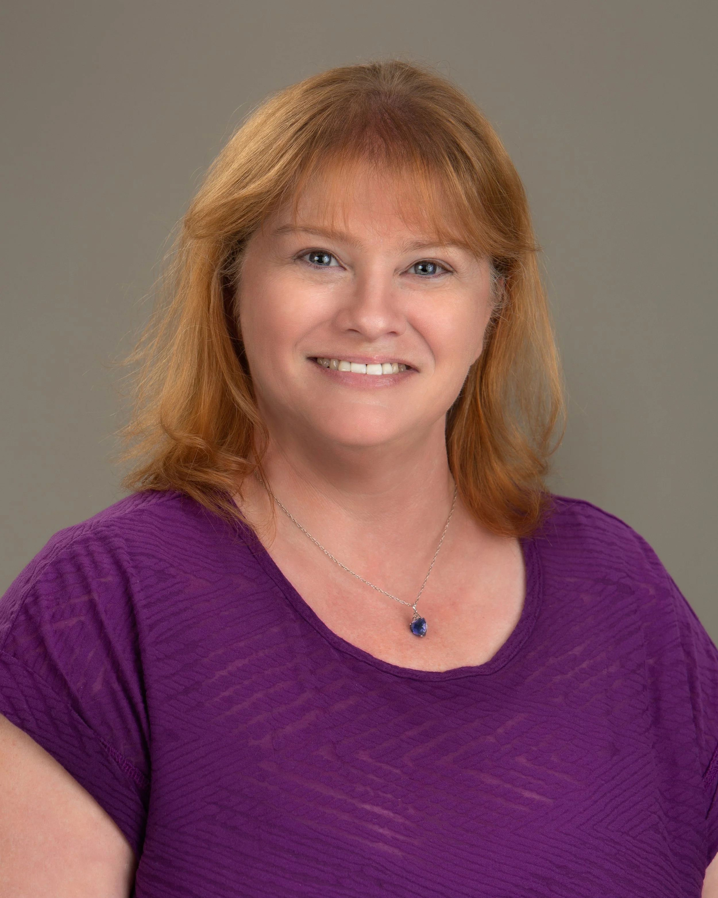 A woman with red hair, wearing a purple top and a necklace with a dark blue pendant, smiling at the camera against a neutral background.
