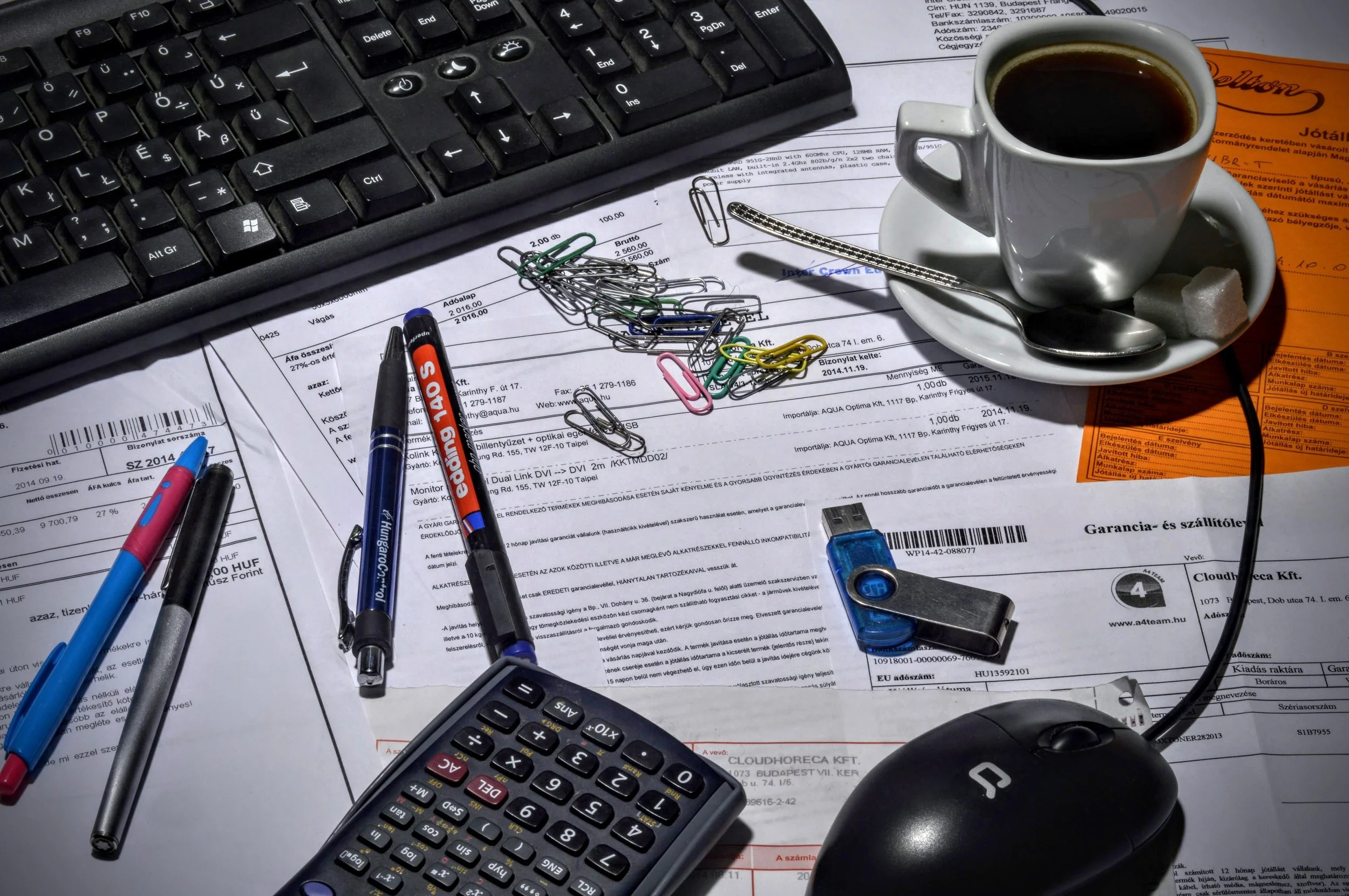 A cluttered workspace with a black computer keyboard, a calculator, a mouse, pens, paper clips, a coffee cup on a saucer, sugar cubes, a pen, and a printed document with text, all arranged on a desk.