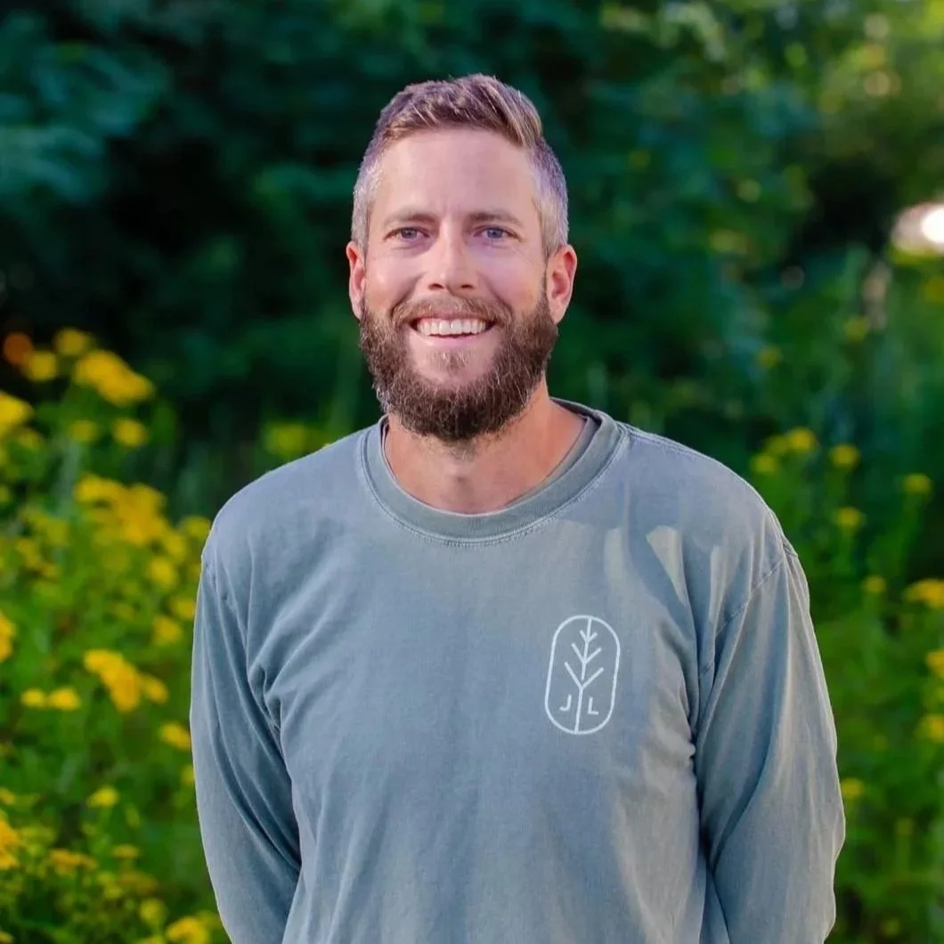A smiling man with a beard and short hair standing outdoors in a natural setting with green foliage and yellow flowers.