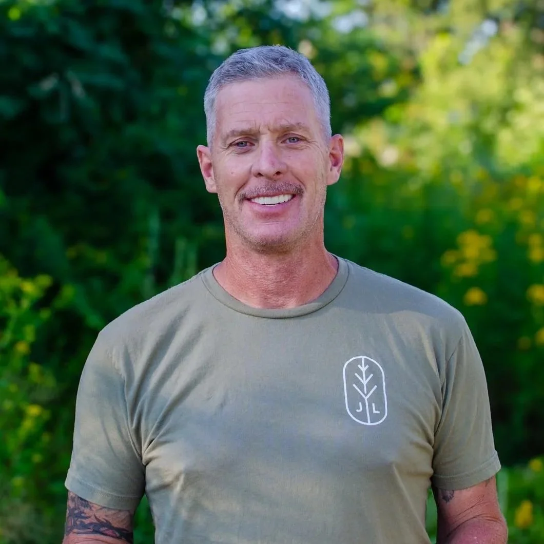 A smiling man with short gray hair standing outdoors with green foliage in the background, wearing a light gray T-shirt with a leaf logo on the chest.
