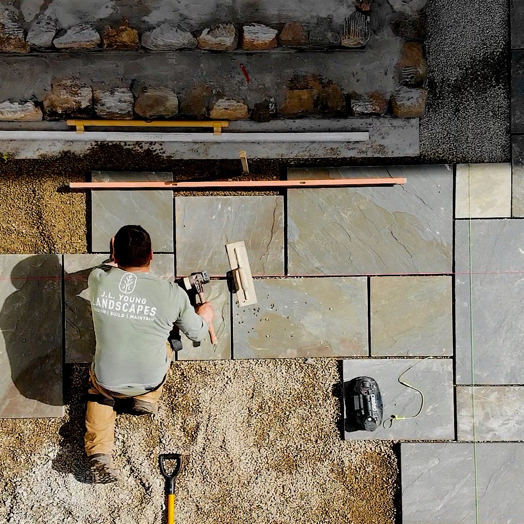 A person installing large stone tiles on a floor with tools, a power drill, and a small radio nearby. The person is gripping a power tool, working on the tiles on a sandy surface, with some stones and construction materials around.