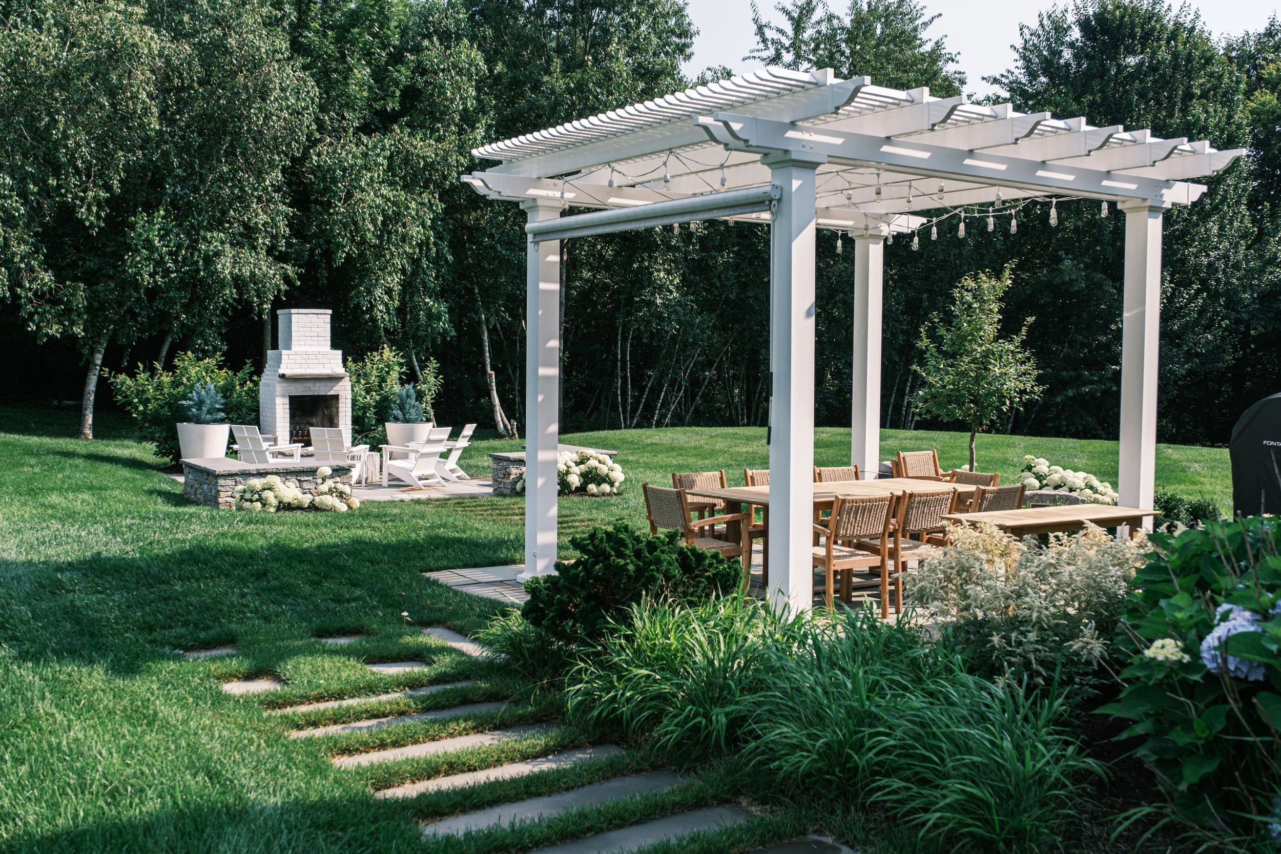 A backyard outdoor patio area with a white pergola, wooden dining table with chairs, a separate seating area with white Adirondack chairs and a fireplace, surrounded by greenery and trees.