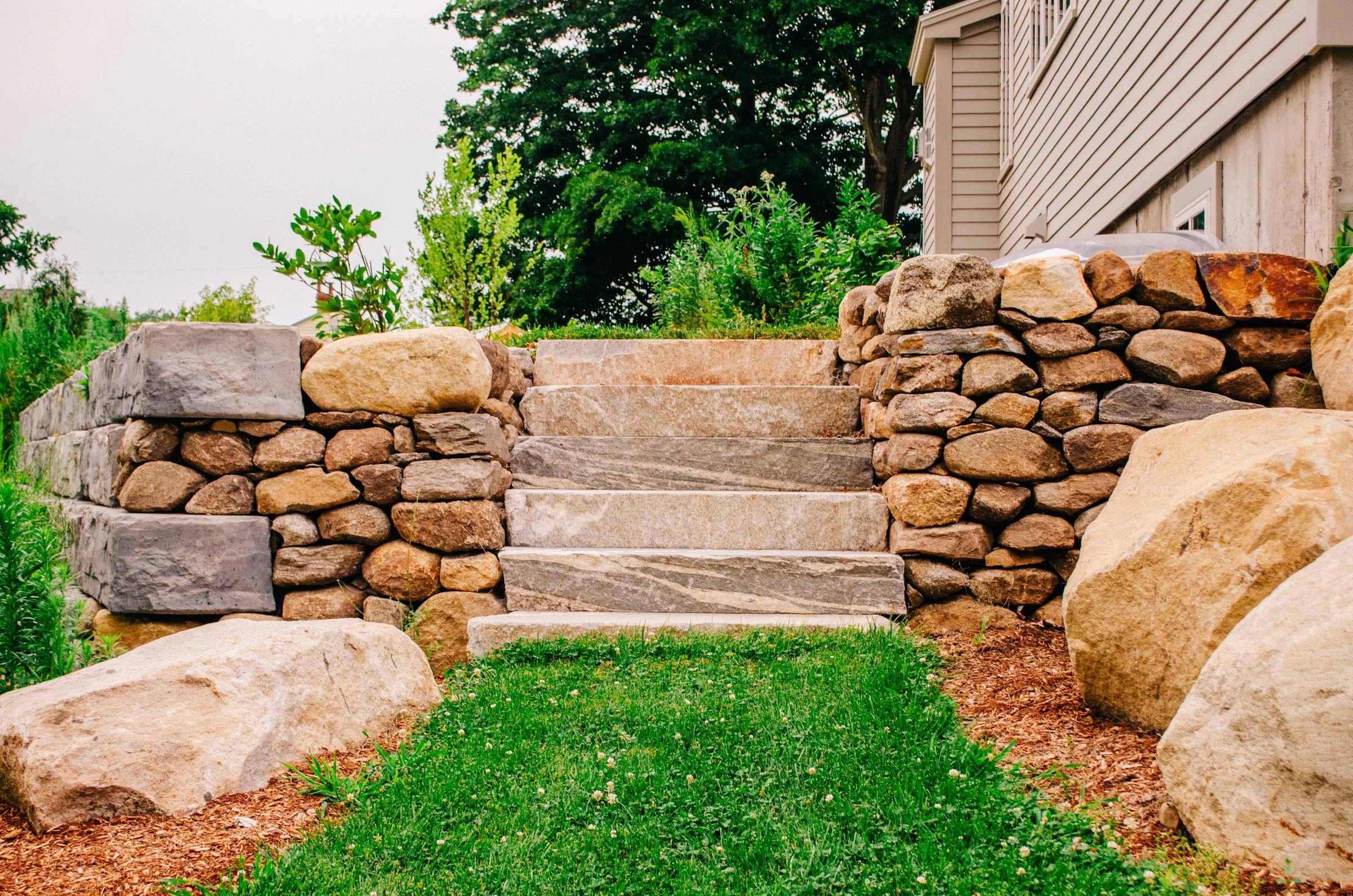Stone steps leading up to a house with beige siding, surrounded by green plants and grass.