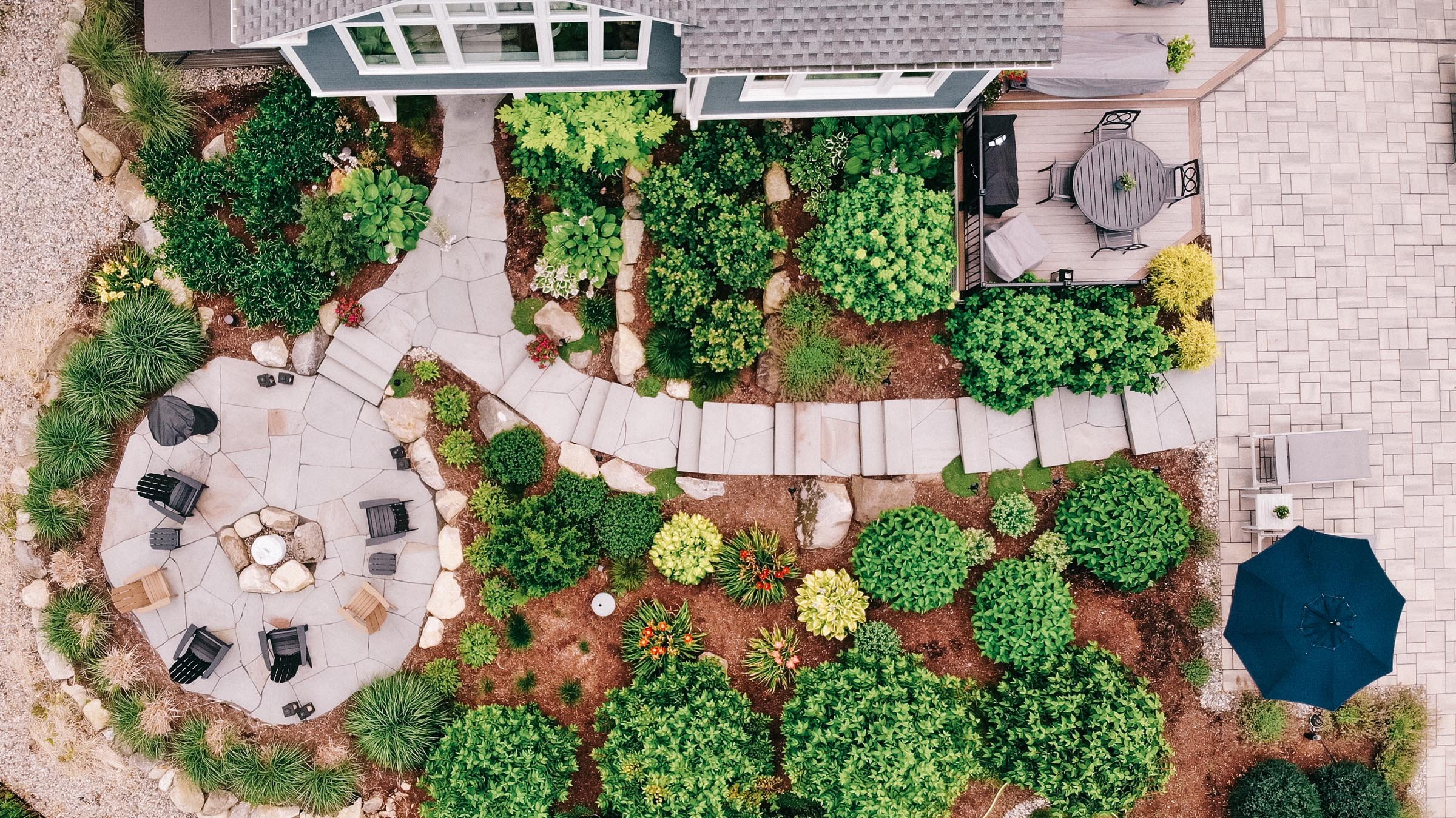 Aerial view of a landscaped backyard with a patio, garden beds with various green plants, rocks, and a small seating area with chairs and a table. There's a wooden deck with outdoor furniture and a blue umbrella. The yard has paved walkways and lush greenery.