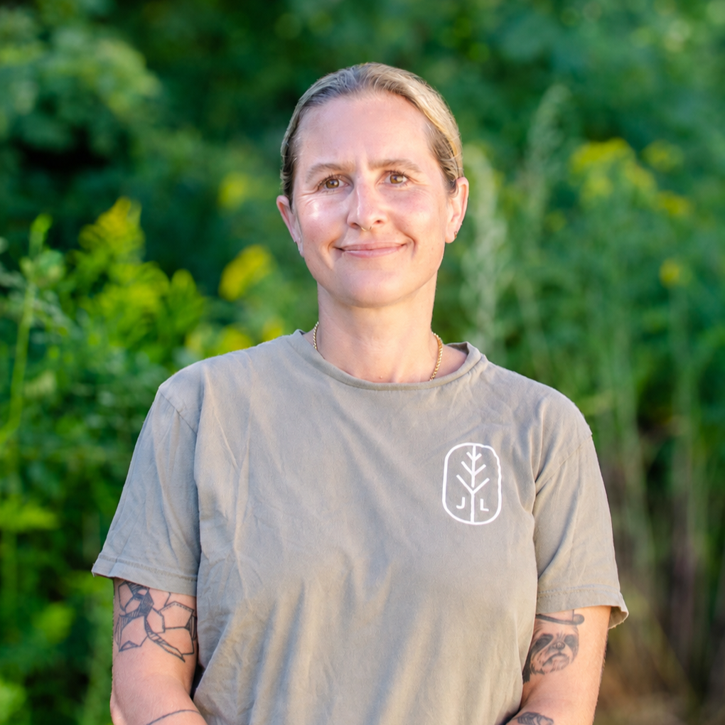 A woman with short blonde hair, wearing a beige t-shirt with a logo on the chest, standing outdoors with green foliage in the background.