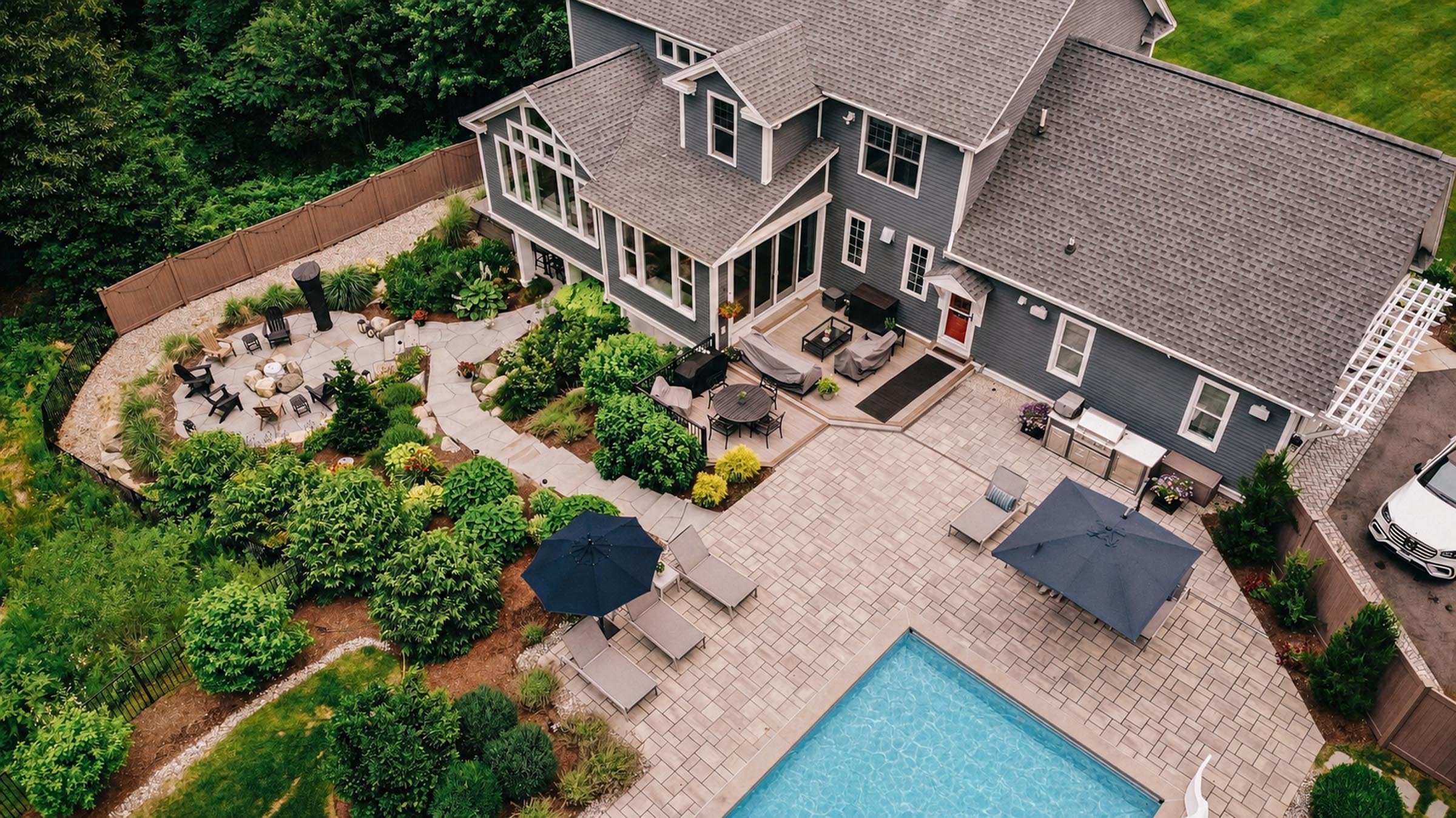 Aerial view of a backyard with a swimming pool, patio, outdoor seating, umbrellas, grill, and landscaped greenery surrounding a modern house.