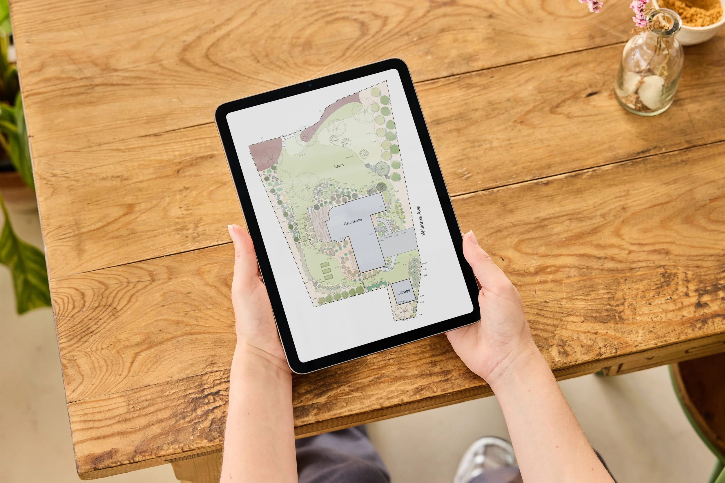 Person holding a tablet displaying a landscape design plan on a wooden table.