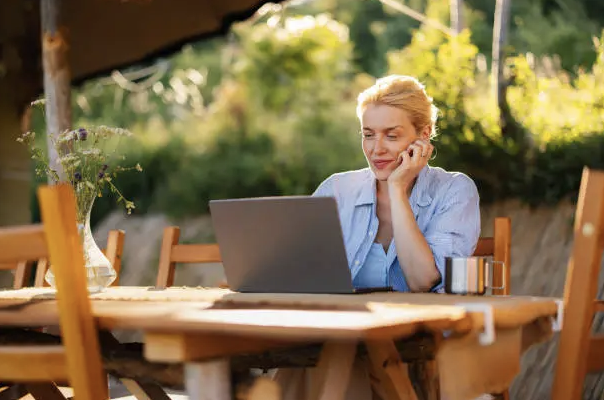 A woman sitting at a wooden table outdoors, working on a laptop with a cup nearby, surrounded by greenery.