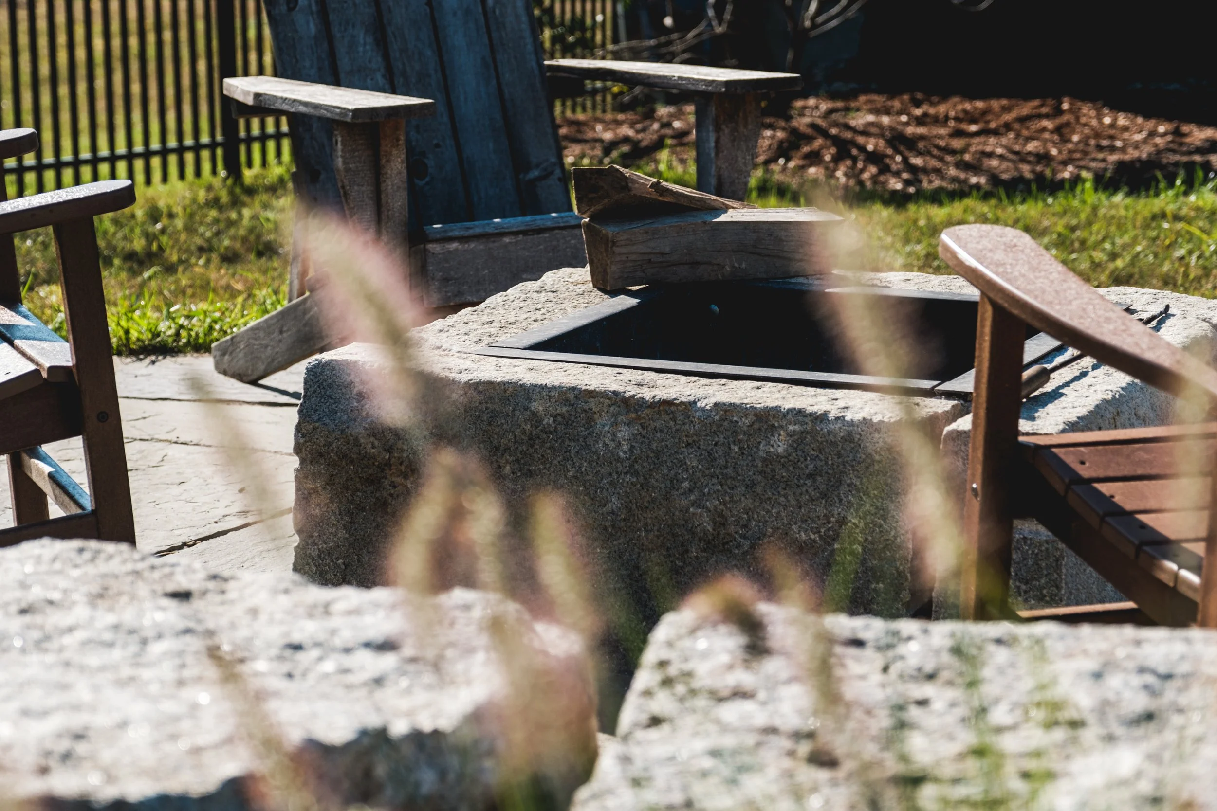 Empty outdoor seating area with two wooden benches and a stone fire pit surrounded by a grassy yard with a black metal fence.
