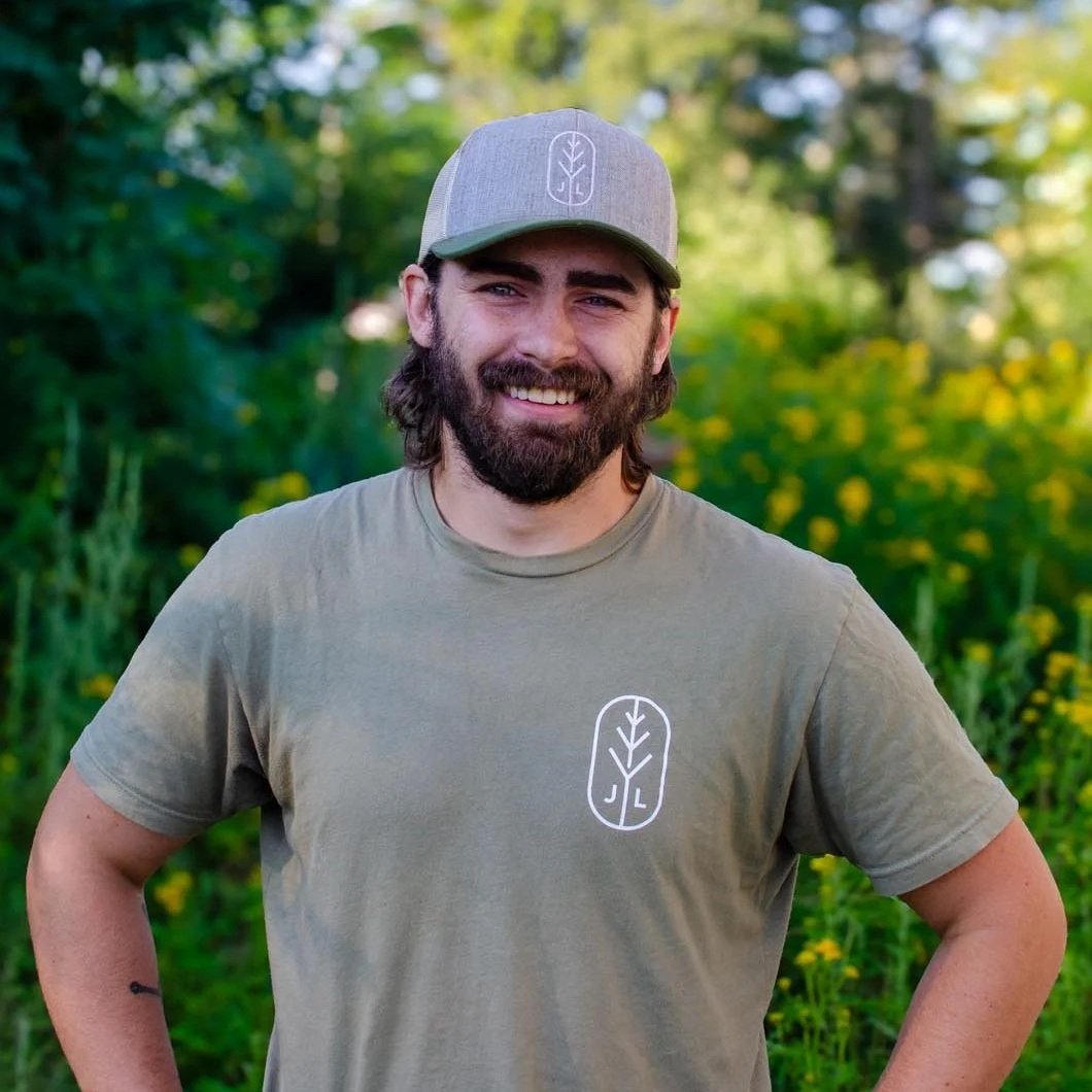 Young man with a beard and long hair wearing a gray baseball cap and a tan T-shirt with a logo of a leaf and initials J L on the chest, standing outdoors in front of green foliage and yellow flowers, smiling.