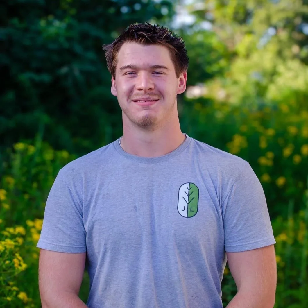 A young man with brown hair and a beard wearing a gray t-shirt with a tree logo, standing outdoors with greenery in the background.