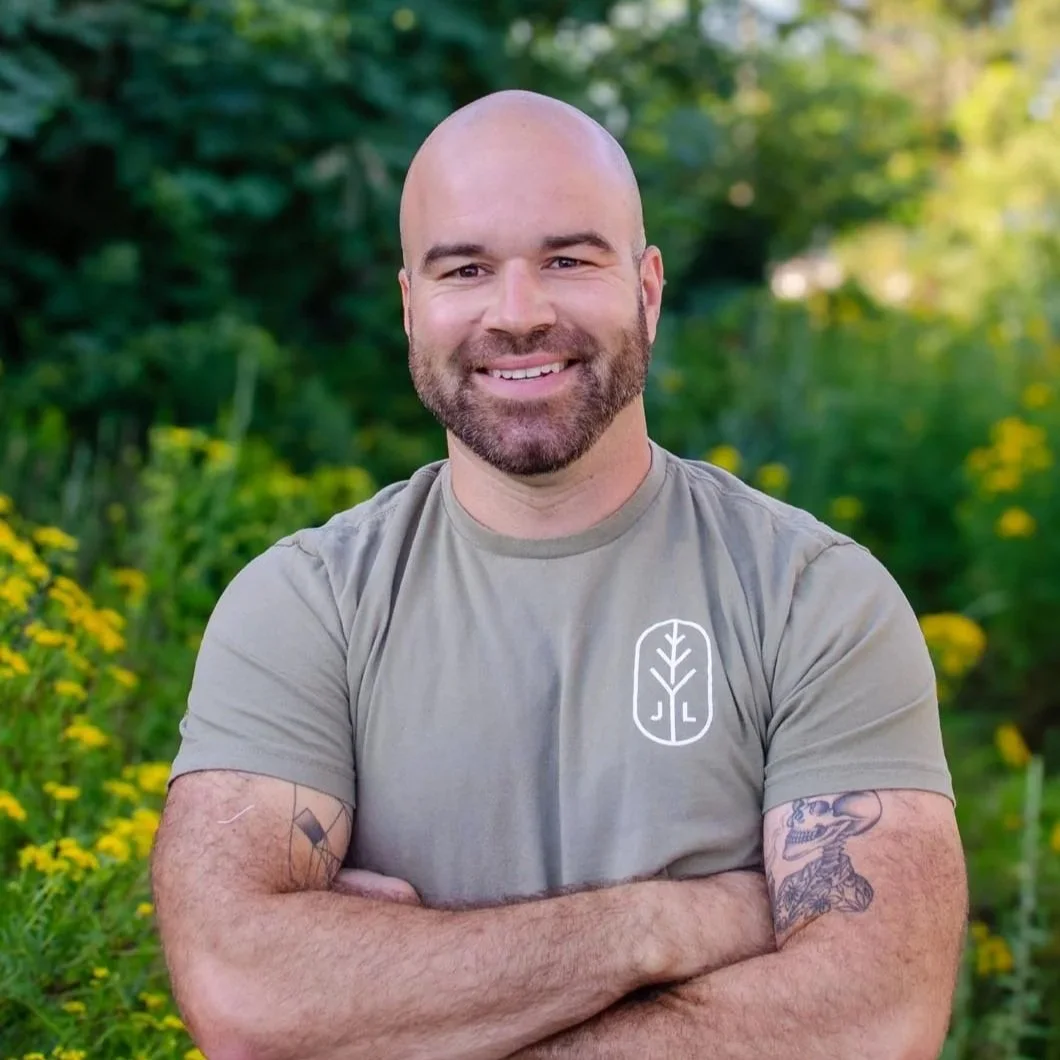 A smiling man with a beard and tattoos on his arms, wearing a light gray t-shirt with a leaf logo, standing outdoors with green foliage and yellow flowers in the background.