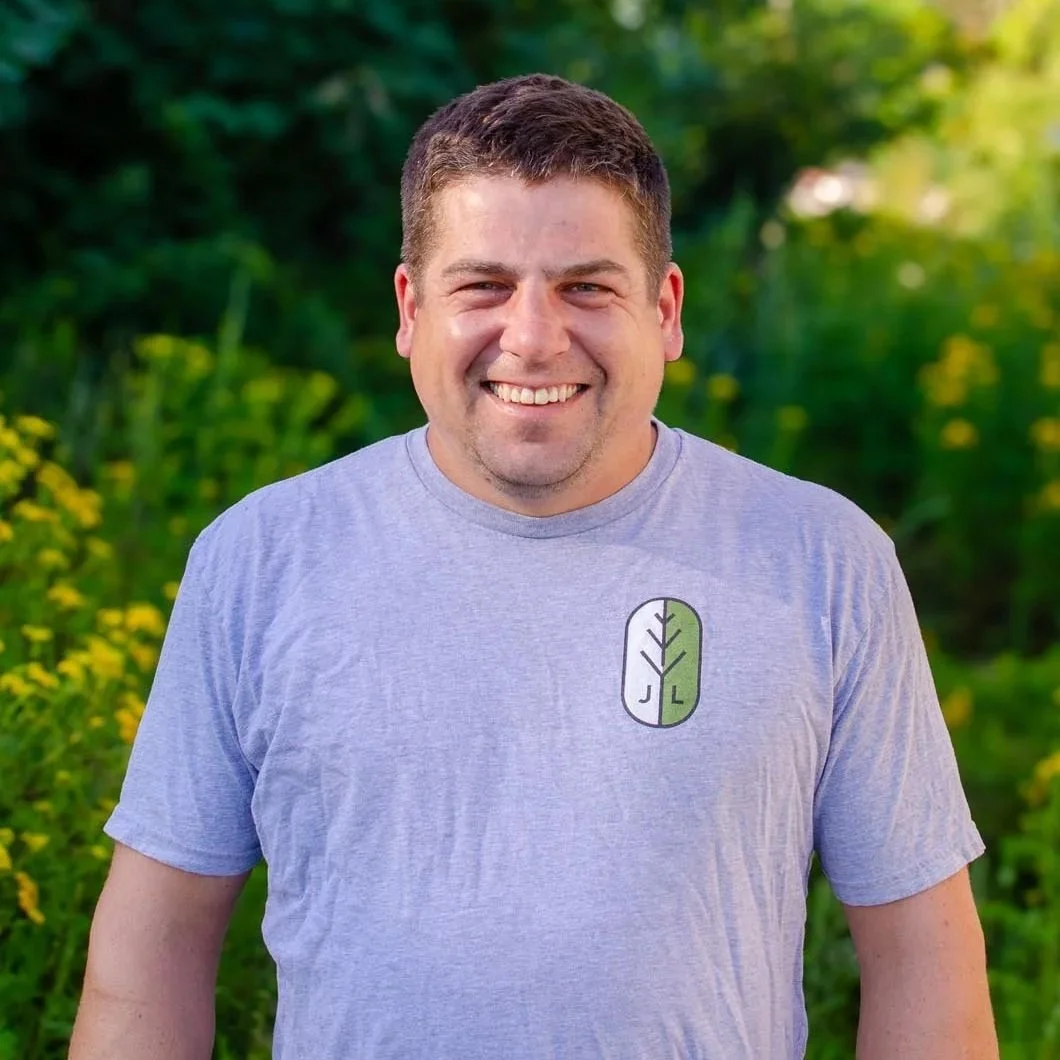 A smiling man in a light gray T-shirt standing outdoors with green foliage and yellow flowers in the background.