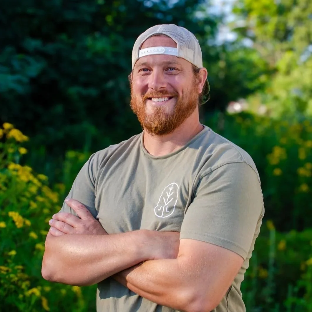 A smiling man with a beard wearing a beige baseball cap backwards and a green T-shirt standing outdoors with green trees and yellow flowers in the background.